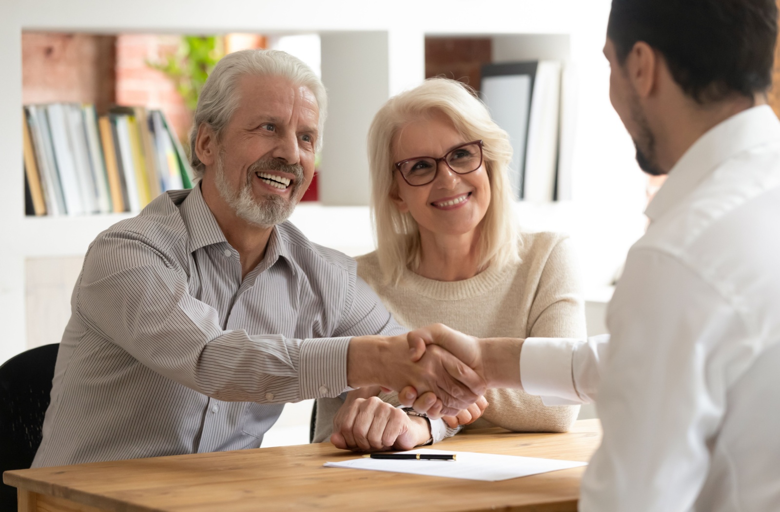 An older adult shakes the hand of a community representative while their smiling spouse watches during an interview with a potential retirement community