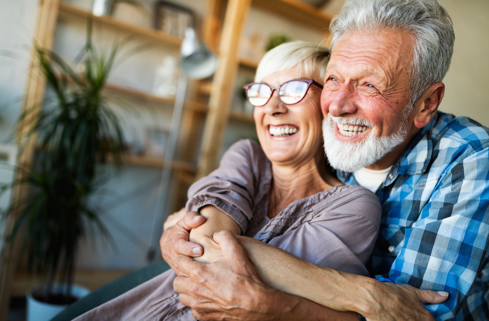 An older adult hugs their laughing spouse from behind while looking out the window in the living room of their new retirement community

