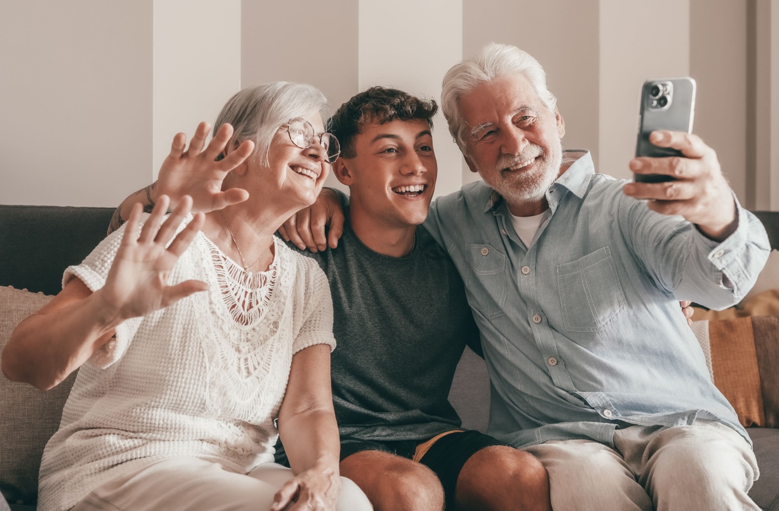 A teenager sits between their grandparents and waves at a cellphone during a video call and a visit