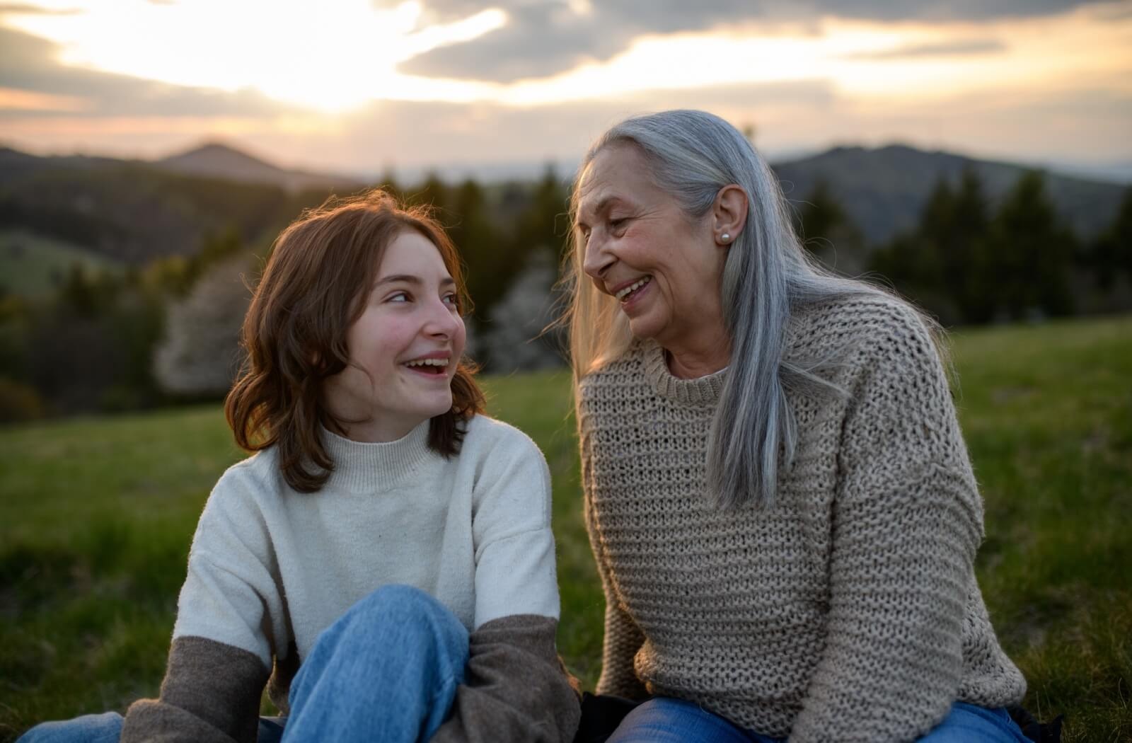 A teenager looks up at their smiling grandparent while sitting on a lush green hill overlooking a forest at sunset