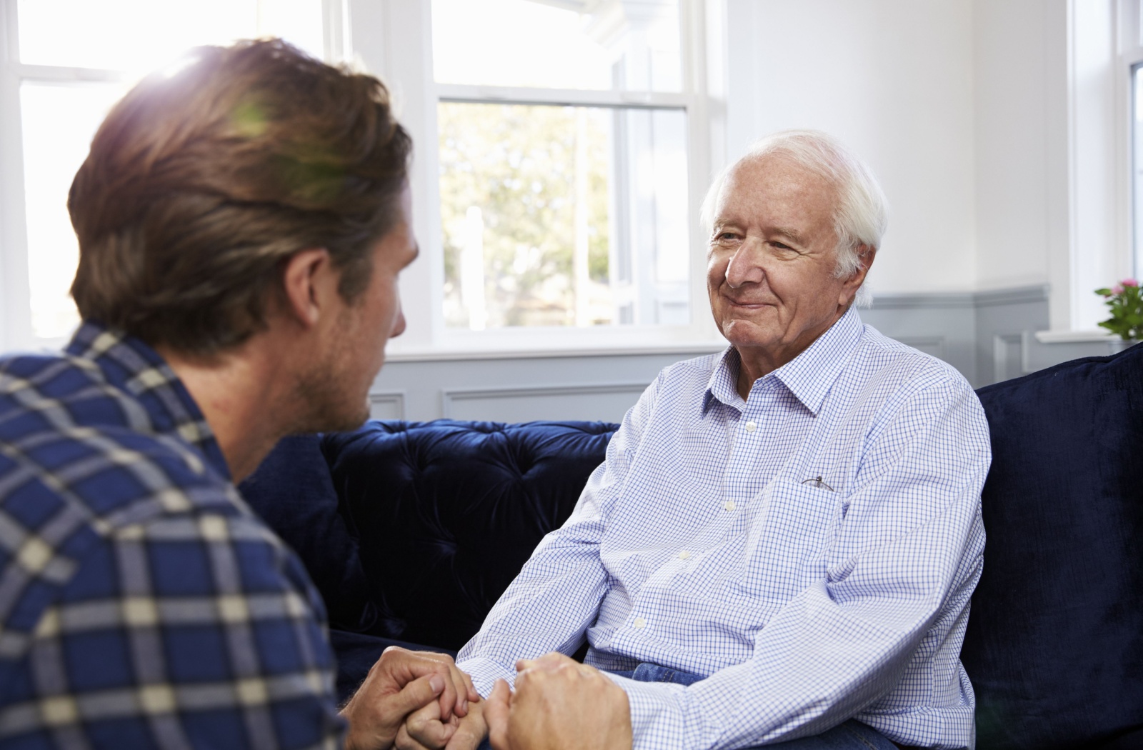 An adult child visiting their senior parent sitting together on a blue sofa holding hands demonstrating care and love