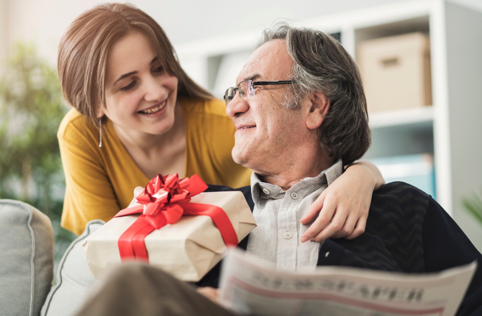 An adult child smiles as they give their senior parent a gift wrapped in a red bow.